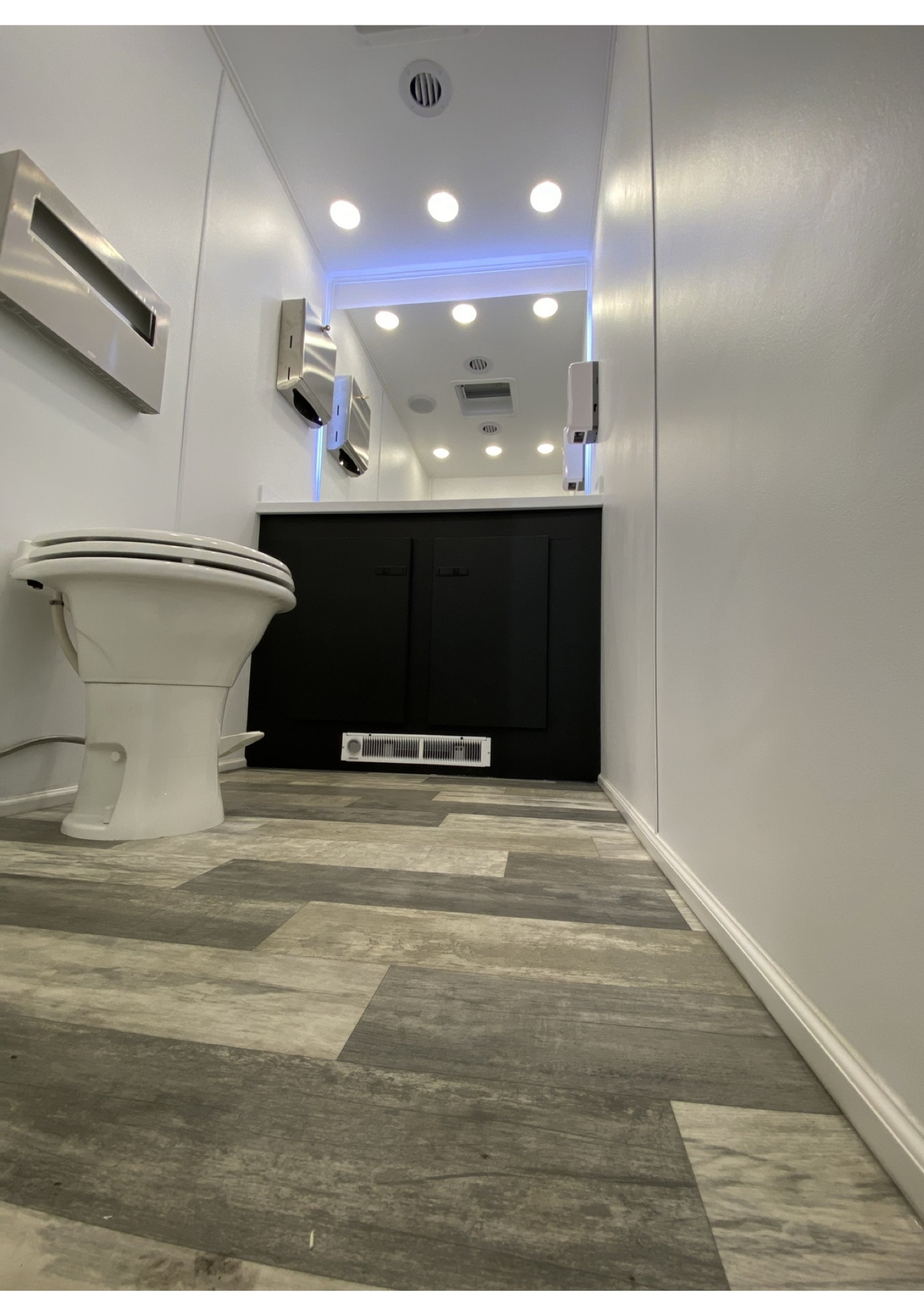 Interior of luxury 2-stall restroom trailer showing vanity with lit mirrors, recessed lighting, and gray plank flooring