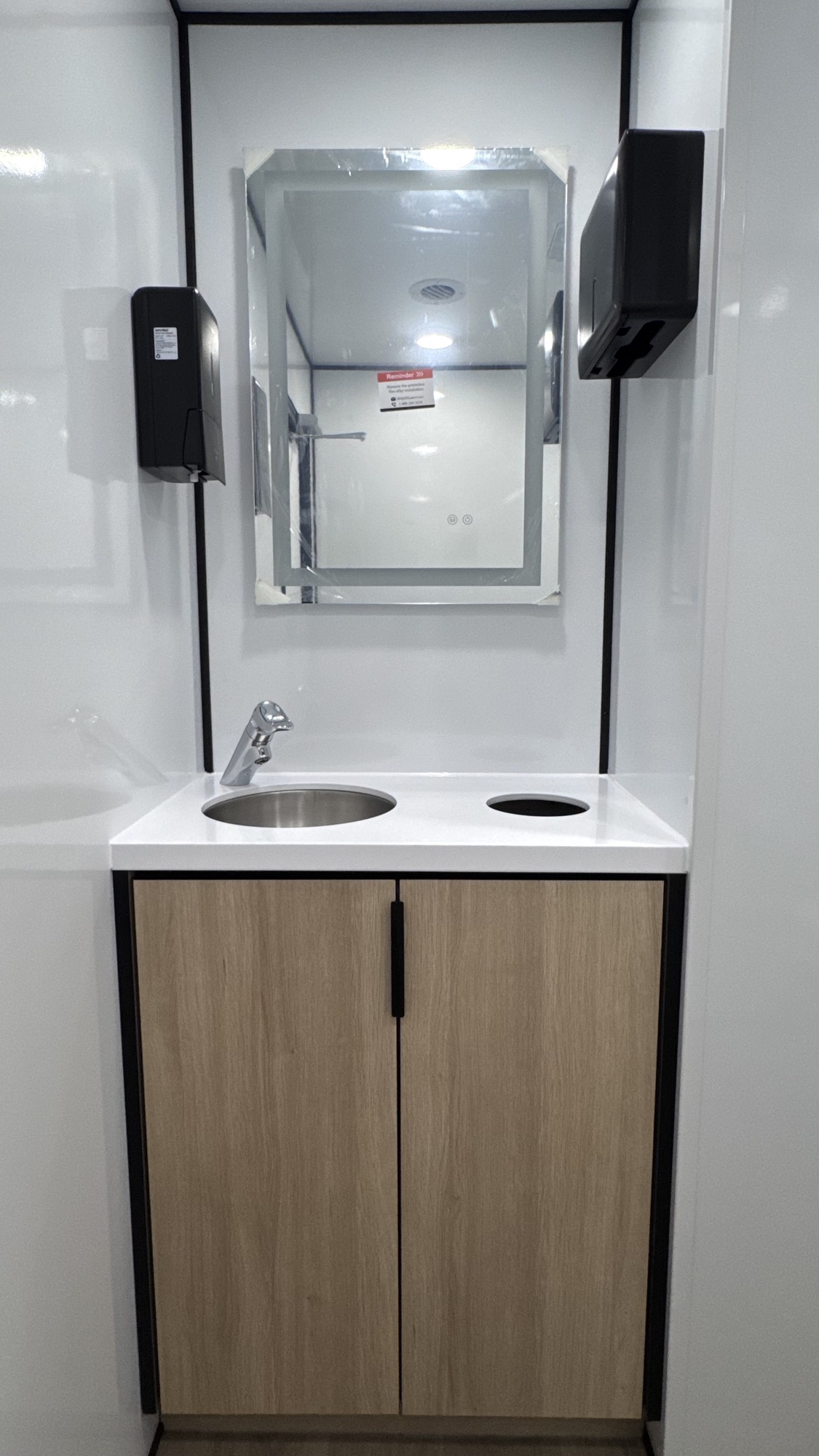 Restroom vanity with stainless basin, chrome faucet, light wood cabinet, framed mirror, and dispensers