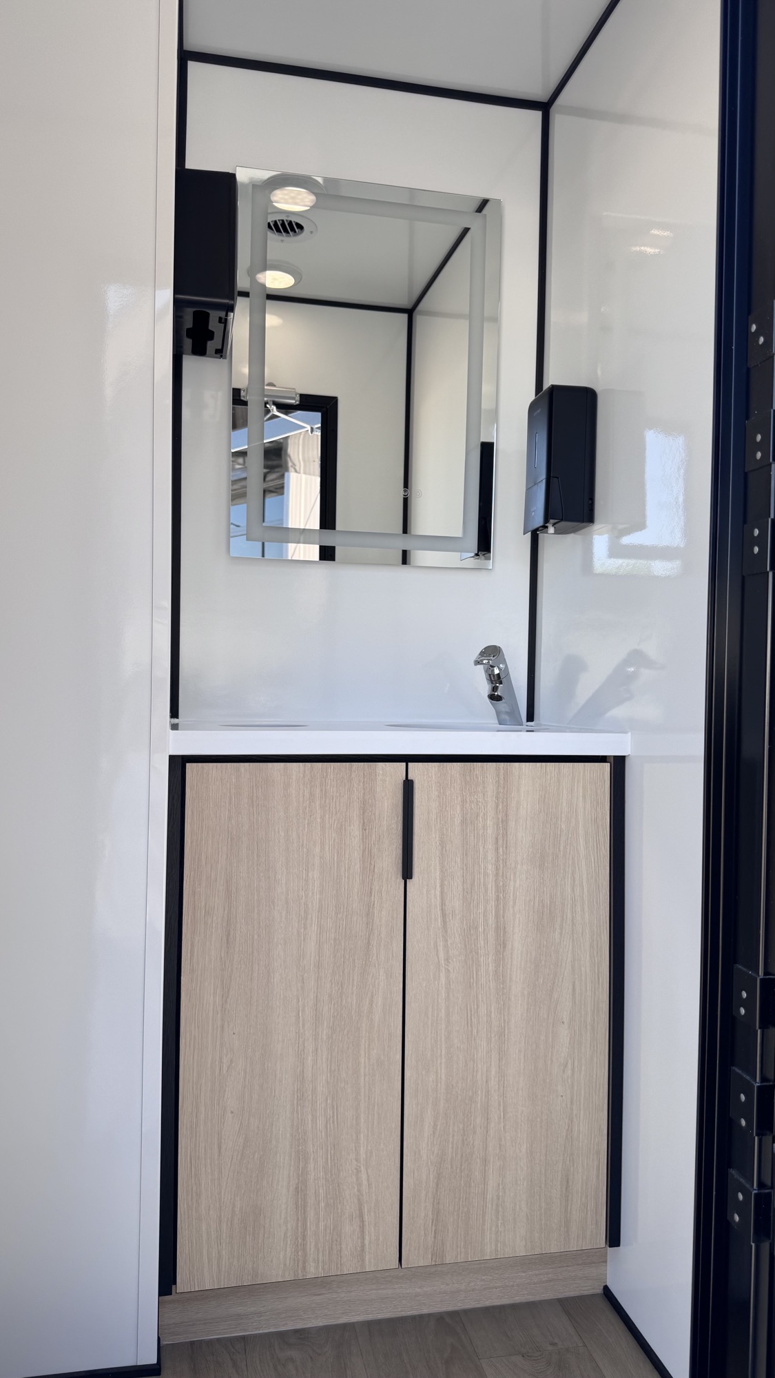 Vanity with backlit mirror, polished chrome faucet, soap dispenser, and wood-grain cabinet inside a private suite